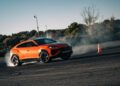 A bright orange Lamborghini Urus SE SUV drifts on an asphalt surface with smoke rising from the tires, past an orange traffic cone. Trees and a clear sky form the backdrop, echoing its thrilling Nardò debut.