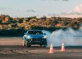 A Lamborghini Urus drifts around orange traffic cones on a paved area, emitting smoke from the tires, with trees and clouds in the background.