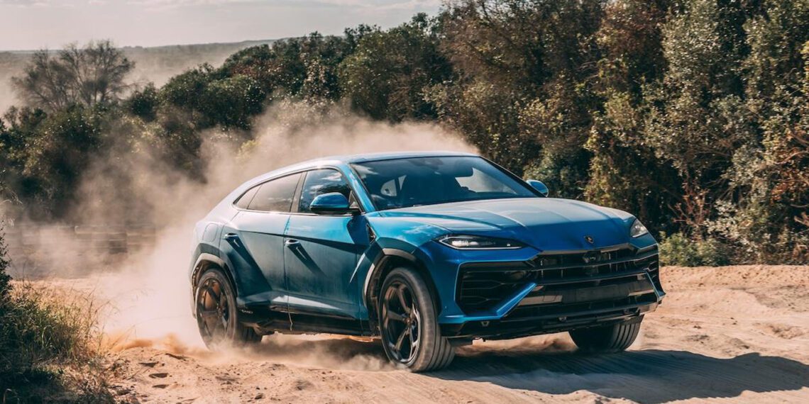 A dynamic debut unfolds as a blue SUV, akin to the hybrid Lamborghini Urus, powers through sandy terrain at Nardò, kicking up clouds of dust against a backdrop of trees and a moody sky.