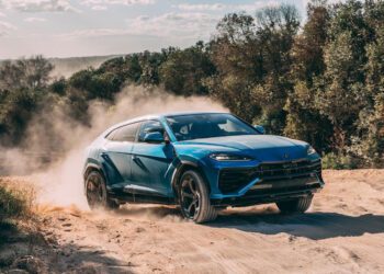 A dynamic debut unfolds as a blue SUV, akin to the hybrid Lamborghini Urus, powers through sandy terrain at Nardò, kicking up clouds of dust against a backdrop of trees and a moody sky.
