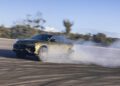 A Lamborghini Urus hybrid drifts on a wide, empty asphalt surface in Nardò, creating a cloud of smoke behind its rear tires. The background features trees and a clear blue sky.