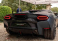 A matte black Lamborghini, a testament to Italy's renowned automotive craftsmanship, boasts distinctive rear lights as it sits gracefully under a canopy on a gravel surface. The scene is quietly complimented by the surrounding greenery, reminiscent of an Autostyle Design Competition showcase.