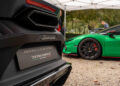 Close-up of a black Lamborghini, flaunting its Italian elegance, with a green sports car parked under a canopy on the gravel surface, reminiscent of an Autostyle Design Competition scene.