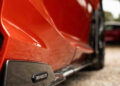 Close-up of a red sports car's side, featuring a "Revuelto" badge, exuding Lamborghini's iconic flair. The car is parked on a gravel surface with blurred greenery in the background, reminiscent of Italy's scenic landscapes.