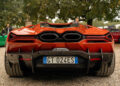 Rear view of a striking red sports car, reminiscent of a Lamborghini, with dual exhausts and a visible license plate, parked on a gravel surface surrounded by picturesque Italian trees.