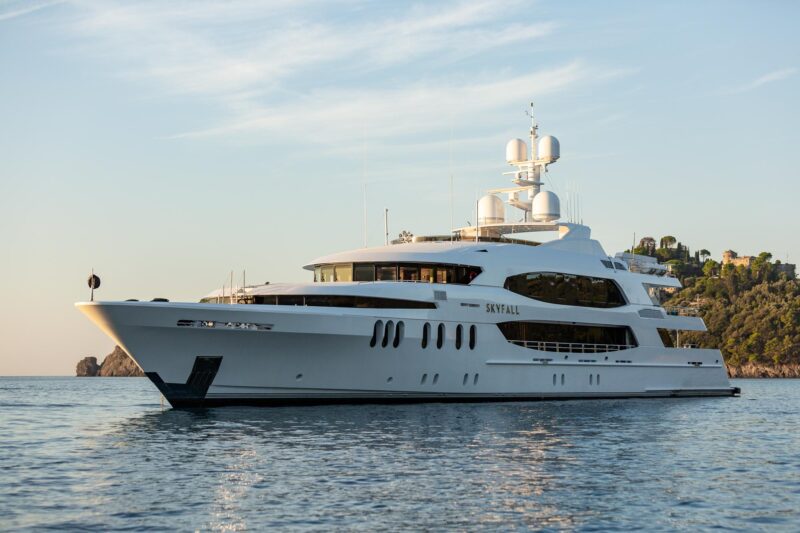 The luxurious white yacht "Skyfall" is anchored on calm waters, framed by a clear sky and hilly coastline. This stunning vessel will be a highlight at the 2024 Fort Lauderdale International Boat Show, showcasing the pinnacle of luxury yachts.