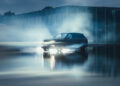 A dark Rolls-Royce Cullinan emits headlights and is surrounded by mist, parked on a reflective wet surface near a glass building.