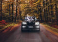 A black Rolls-Royce Cullinan glides down a tree-lined road, surrounded by blurred autumn foliage, exuding luxury and custom sophistication.