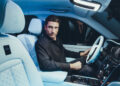 A man seated in the driver's seat of a Rolls-Royce Cullinan, holding the steering wheel, enjoys the modern interior featuring ambient lighting and detailed upholstery.