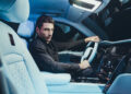 A person sits in the driver's seat of a Rolls-Royce Cullinan with soft blue interior lighting, holding the steering wheel and looking toward the camera.