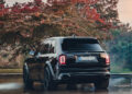 A black luxury SUV, customized to perfection, is parked on a reflective surface, with a tree displaying autumn foliage in the background.