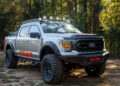 A silver off-road truck with large tires and roof lights, parked on a dirt path. The word "Bear" is emblazoned in red on the hood and sides. Trees stand tall in the background, creating a perfect scene for any Adventure Garage enthusiast.