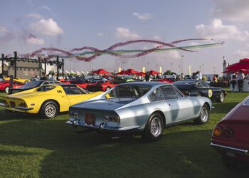 Vintage cars, including a stunning Ferrari, are displayed on a grassy field with colorful smoke trails painting the sky. Enthusiasts gather under red umbrellas in the background, celebrating the legacy of Enzo Mattiolo and Cavallino Inc.'s passion for automotive excellence.