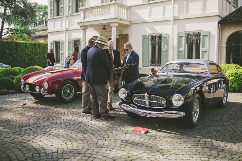 People gather beside two vintage cars from Cavallino Inc., displayed on a cobblestone driveway, with a classic white building in the background.