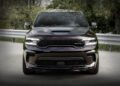 Front view of a dark-colored Dodge Durango SRT with a sleek design, parked on a roadway. The SUV's imposing stance and HEMI Power are highlighted by the surrounding trees and metal guardrails.