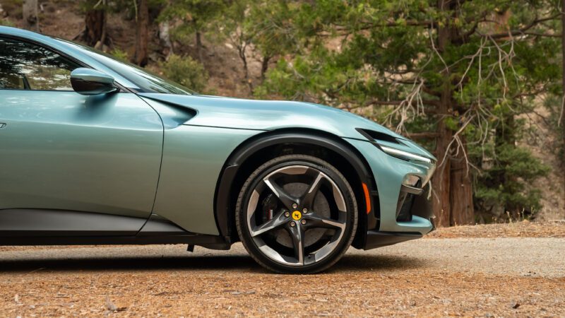 Close-up of the front side of a sleek turquoise sports car on a dirt road with trees in the background.