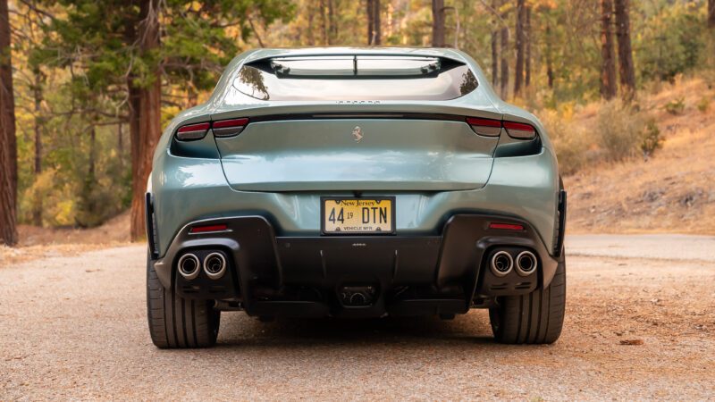 A green Ferrari sports car parked on a forest road, viewed from the rear, with a visible license plate. Trees and foliage surround the scene.