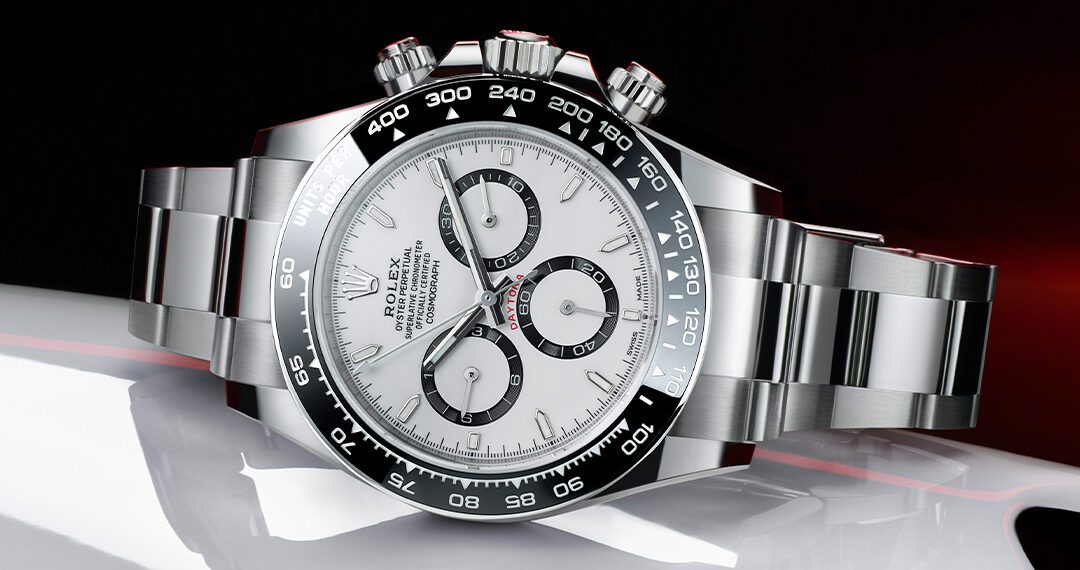 A stainless steel wristwatch with a white dial and black bezel, featuring three subdials and a chronograph function, is showcased as part of the Timeless Treasures collection. Positioned on a reflective surface against a dark background, it exudes elegance reminiscent of Monterey Car Week's sophistication.