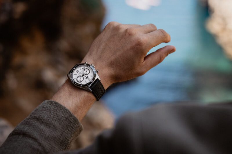 A person wearing a Glashütte Original SeaQ Chronograph with a silver face and black strap, standing outdoors near a blurred body of water.