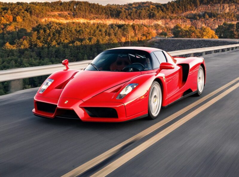 A sleek 2003 Ferrari in Rosso Corsa cruises down the road, with lush trees painting a vivid background.