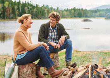 A man and woman sit on logs by a lakeside, smiling and talking. Clad in sweaters and jeans from the latest Fall Shopping Guide, they enjoy the cozy ambiance of a small campfire. A picturesque forest and majestic mountains provide the perfect backdrop for this serene moment.