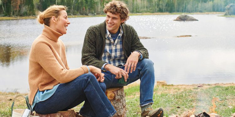 A man and woman sit on logs by a lakeside, smiling and talking. Clad in sweaters and jeans from the latest Fall Shopping Guide, they enjoy the cozy ambiance of a small campfire. A picturesque forest and majestic mountains provide the perfect backdrop for this serene moment.