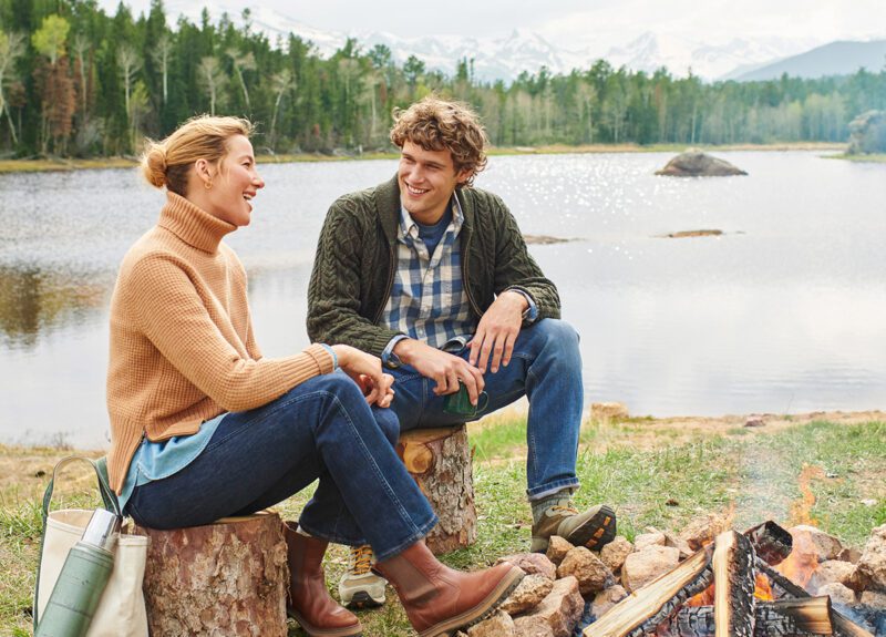 A man and woman sit on logs by a lakeside, smiling and talking. Clad in sweaters and jeans from the latest Fall Shopping Guide, they enjoy the cozy ambiance of a small campfire. A picturesque forest and majestic mountains provide the perfect backdrop for this serene moment.
