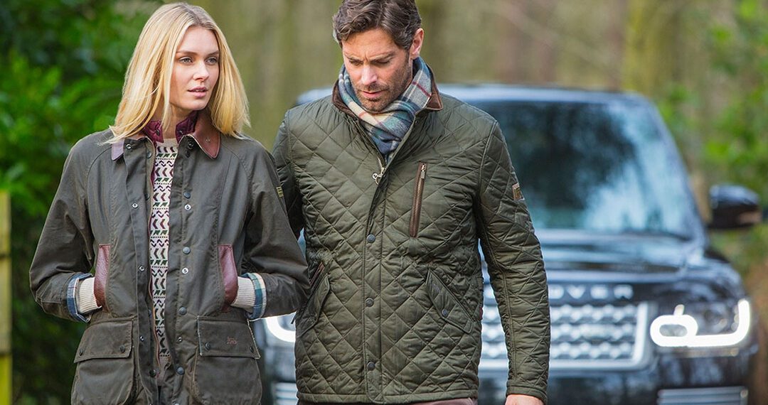 A man and woman stroll outdoors in quilted jackets, showcasing Lux Styles, with a dark SUV from the duPont REGISTRY parked behind them.