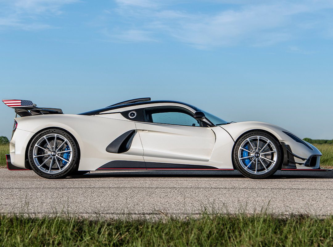 A sleek white sports car, reminiscent of the finest American supercars, with a rear spoiler and blue accents, is parked on grass, viewed from the side.