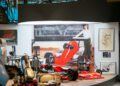 The museum exhibit showcases a striking red race car, complemented by authentic engine parts and a large black and white photograph of a person standing proudly next to the Pagani.