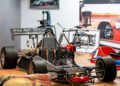 A display of a vintage race car chassis, a bicycle, and racing memorabilia in a museum setting, featuring the iconic engineering of the Utopia Roadster.