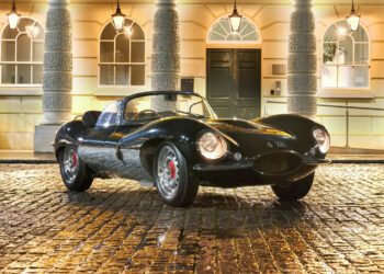 A vintage 1957 Jaguar XKSS sports car is parked on a wet cobblestone street, with an elegant building in the background, its allure capturing the essence of classic luxury—an icon that once fetched $14 million at an RM Sotheby's Auction.