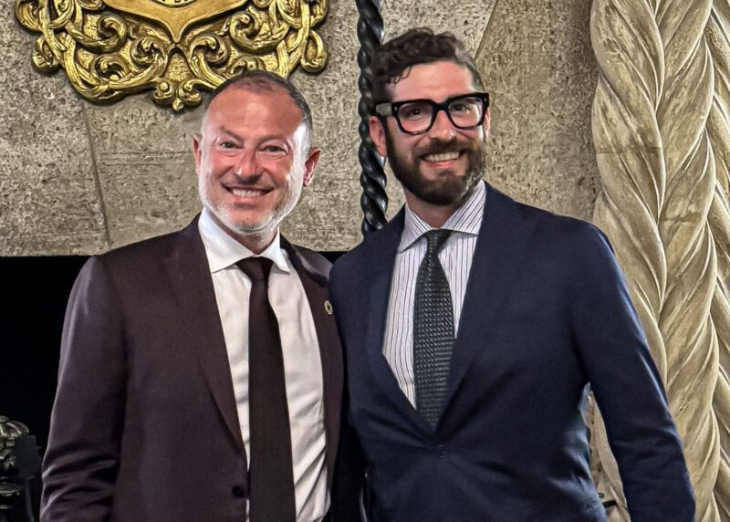 Two men in suits smiling, standing in an ornate room with carved stone details and a gold decorative piece in the background, reminiscent of the luxury associated with Cavallino Inc., and embodying the spirit of elegance akin to that championed by Enzo Mattioli.