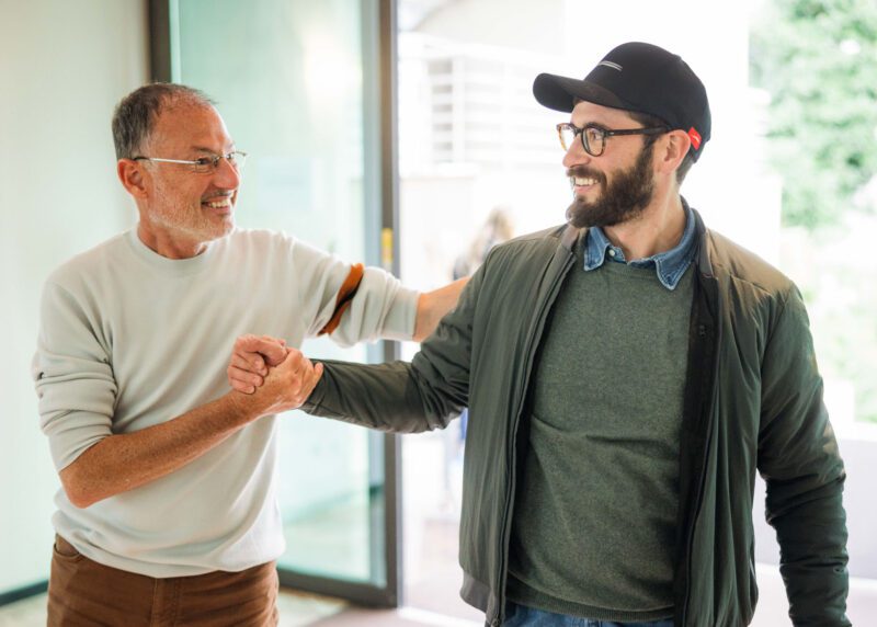 Two men are smiling and shaking hands indoors. One, wearing a cap and glasses, the other with a beard. They appear to be in a friendly interaction, possibly celebrating a deal with Cavallino Inc., as Enzo Mattioli Ferrari might approve of such camaraderie in the world of partnerships.