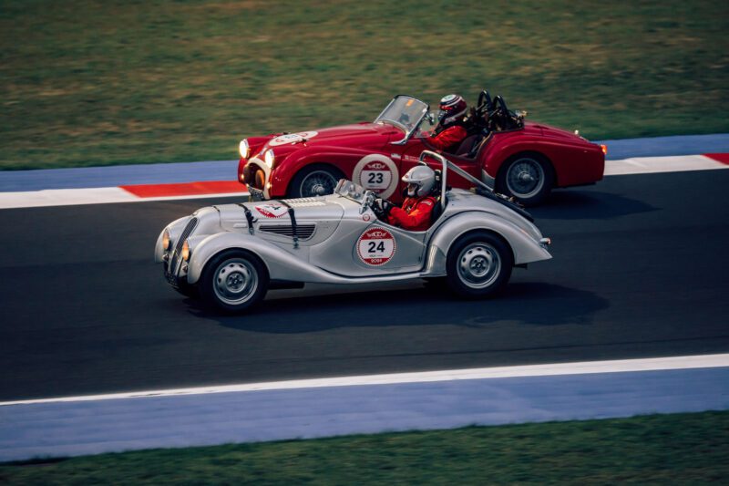 In a thrilling recap of the 2024 Modena Cento Ore, two vintage race cars, one silver and one red, with drivers in helmets, race side by side on a track with grass flanking either side.