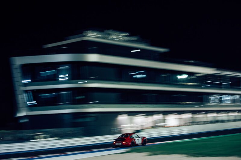 A red car, reminiscent of the thrilling Cento Ore races, speeds past a blurred multi-story building on a racetrack under the night sky.