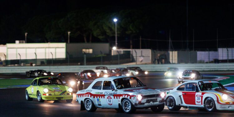 A group of vintage race cars competes on a dimly lit track during the Modena Cento Ore night race, with blurred trees and buildings in the background.