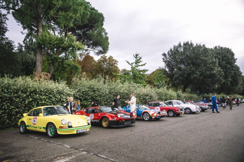 A row of vintage cars, reminiscent of the Modena Cento Ore, is parked outdoors on an overcast day, with people standing nearby. Trees and bushes form the background.