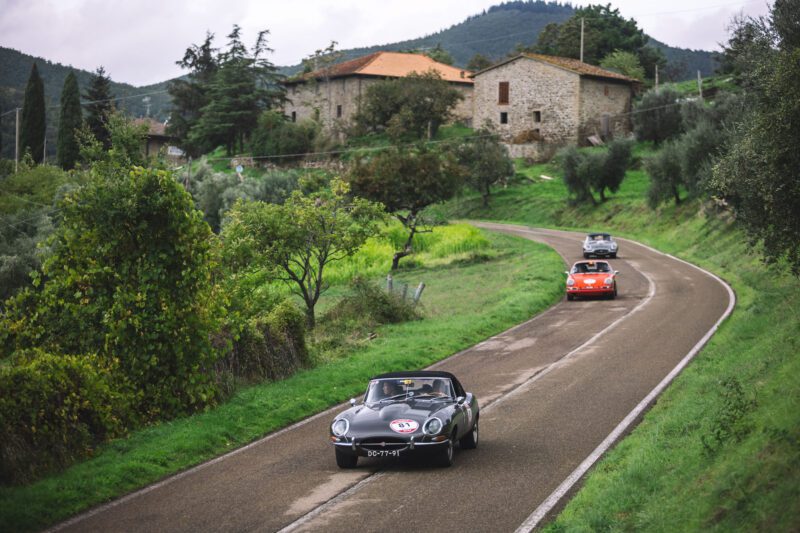 In a recap of the exhilarating 2024 Modena Cento Ore, classic cars glide along a scenic, winding rural road, enveloped by lush greenery and historic stone buildings in the background.