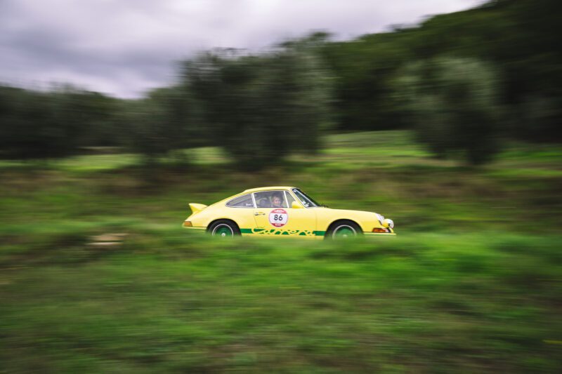 A yellow vintage race car with number 86 on the side speeds through a grassy area with blurred motion, reminiscent of the thrilling races at the Modena Cento Ore in a stunning homage to classic motorsports.