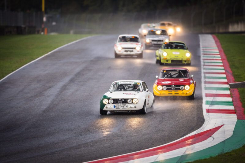 In a thrilling recap of the 2024 Modena Cento Ore, multiple vintage cars race on a wet track, with two leading cars closely competing.