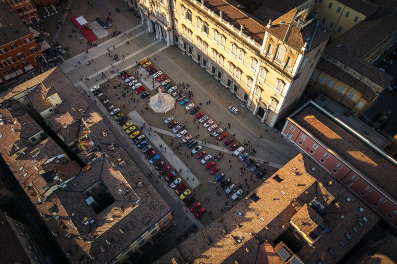 Aerial view of a bustling plaza during the Modena Cento Ore, showcasing rows of colorful parked cars and people mingling. Surrounded by historic buildings with terracotta roofs, this picturesque scene captures the event's vibrant atmosphere.