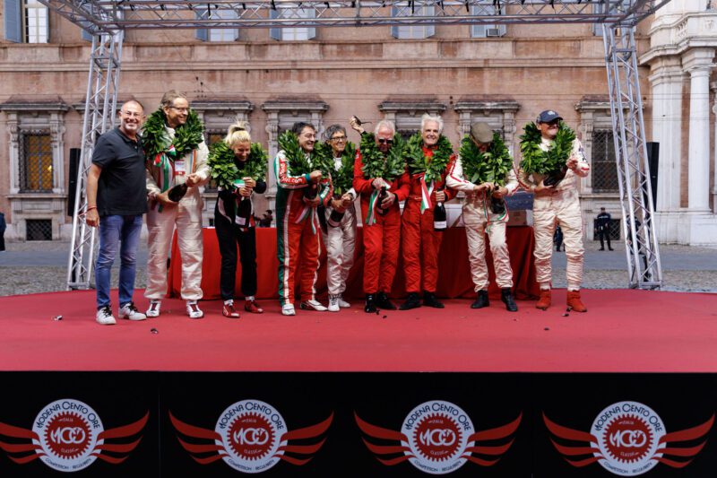 A group of people in racing suits stands proudly on a red podium adorned with laurel wreaths, capturing the triumph of the Modena Cento Ore 2024, set against the backdrop of a historic building.