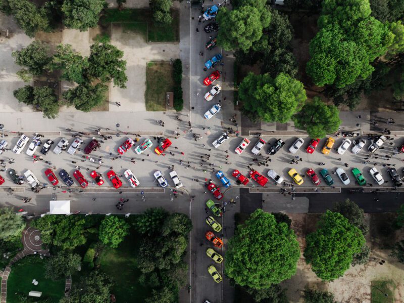 An aerial view of the Modena Cento Ore car show reveals vibrant, colorful cars parked in a tree-lined setting. The pathways weave through lush greenery as attendees stroll around, making it a picturesque 2024 recap of automotive elegance.