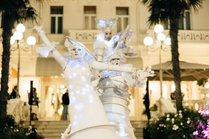 Three performers in elaborate, white, futuristic costumes pose dramatically in front of an ornate building with warm lighting and decorative columns, reminiscent of a scene from the Modena Cento Ore.