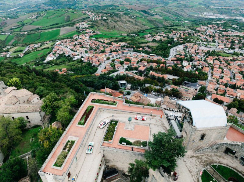 Aerial view of a historic fortress with stone buildings and a courtyard, surrounded by greenery. It overlooks a sprawling town with red-roofed houses and rural landscape in the background, reminiscent of the timeless allure seen during events like the Modena Cento Ore 2024.