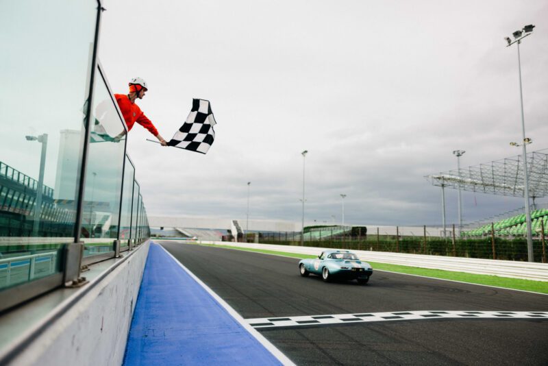 As the race car roars across the finish line, an official waves a checkered flag on the storied racetrack, reminiscent of a thrilling Modena Cento Ore.