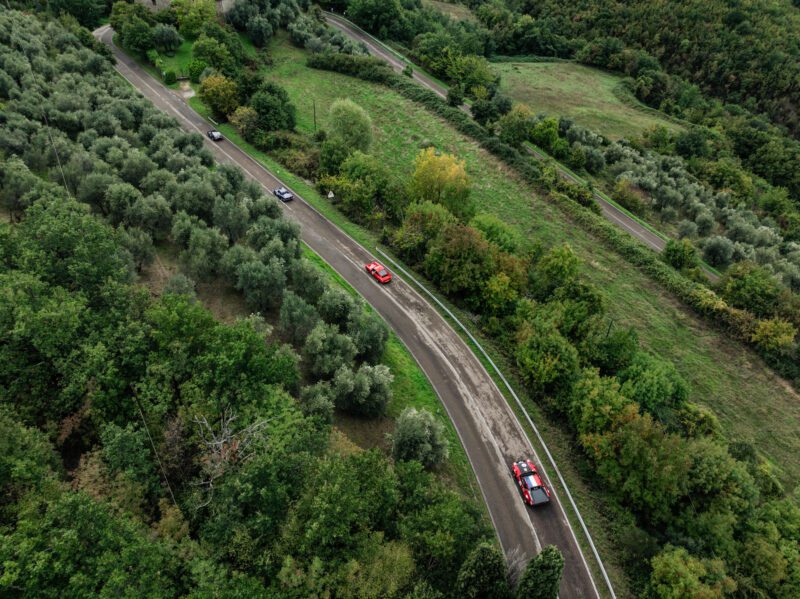 Aerial view of a winding road through a lush green landscape, with several red and black cars driving along it, reminiscent of the iconic Modena Cento Ore race.