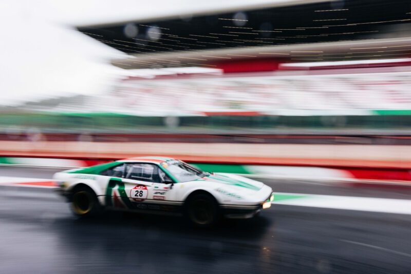 In the 2024 Modena Cento Ore, race car number 28 speeds on a wet track, creating motion blur against a blurred grandstand backdrop.