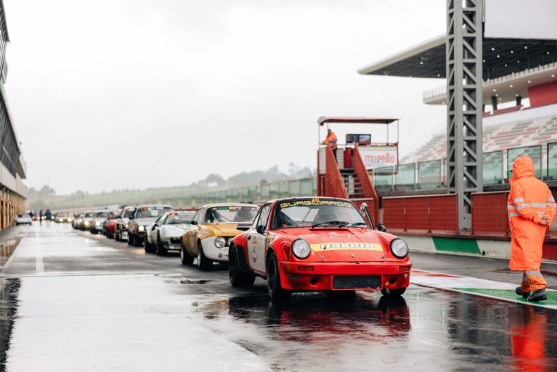 A lineup of classic race cars waits on a wet track at Mugello Circuit during the Modena Cento Ore 2024. A person in an orange rain suit stands nearby, capturing the essence of this iconic event.